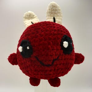 Pode incluir: Um brinquedo de pelúcia vermelho de crochê com orelhas brancas e olhos pretos. O brinquedo tem um rosto sorridente e pequenos braços e pernas.