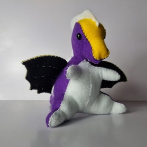Puede incluir: Un dragón de fieltro morado y blanco con alas de fieltro negro. El dragón tiene una cresta de fieltro amarillo y un ojo negro.