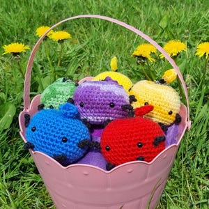 Puede incluir: Una cesta rosa llena de seis animales de peluche tejidos a crochet. Los animales son de varios colores, incluyendo morado, azul, verde, amarillo y rojo. La cesta está sentada sobre un lecho de hierba verde.