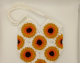May include: A white crochet bag with a sunflower pattern. The bag features a series of crocheted squares, each depicting a sunflower with orange and yellow petals and a brown center. The bag has a white strap.