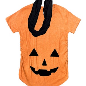 May include: Orange t-shirt featuring a black jack-o'-lantern face design. A black infinity scarf is draped over the shirt. The shirt has short sleeves and a curved hemline, ideal for Halloween.