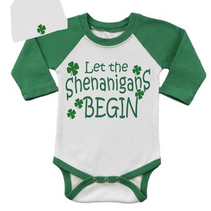 May include: White baby onesie with green sleeves and a green trim. The onesie has the text "Let the Shenanigans Begin" with shamrock designs. A white beanie with a green shamrock design is included.