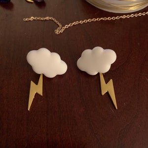 May include: A pair of white cloud earrings with gold lightning bolt pendants. The earrings are set on a dark brown surface, with other jewellery and gold wire visible in the background.