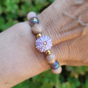 May include: A beaded bracelet featuring a central lavender sea urchin bead, flanked by light brown and iridescent beads. The bracelet includes gold-colored spacers and light pink beads, creating a unique and colorful accessory.