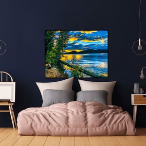 May include: A bedroom interior with a bed, a nightstand, and a framed landscape painting of a lake with a sunset sky. The painting is above the bed, which has a pink comforter and gray pillows. The nightstand is on the right side of the image and has a lamp and a vase on it.