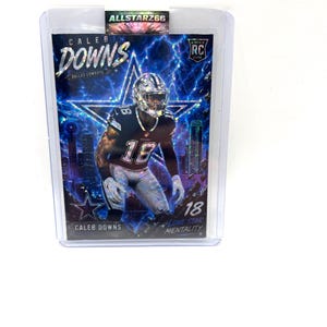May include: A Caleb Downs Dallas Cowboys football rookie card in a protective sleeve. The card features a player in a helmet and uniform, with the number 18. The background has a star and city skyline design. The text "Lone Star Mentality" is visible.