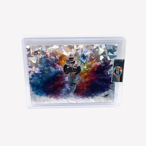 May include: A rectangular trading card featuring a football player, De'Von Achane, in a white uniform. The background displays a vibrant, multi-colored explosion of smoke and a geometric, shattered glass pattern. The card is encased in a clear plastic holder.