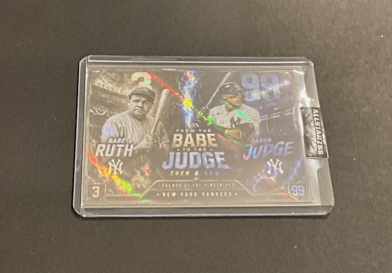May include: A baseball trading card featuring Babe Ruth and Aaron Judge. The card has a holographic design with the text "From the Babe to the Judge" and "Legends of the Pinstripes". The card is encased in a protective sleeve.