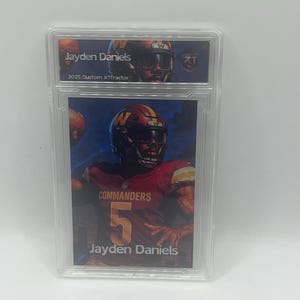 May include: A graded trading card featuring Jayden Daniels in a Commanders uniform. The card is encased in a clear plastic holder. The card has the text "Jayden Daniels" and "2025 Custom XTFractor". The player is wearing a burgundy and gold uniform.
