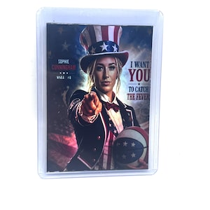 May include: A rectangular sports trading card in a clear plastic sleeve featuring Sophie Cunningham dressed as Uncle Sam in a red, white, and blue top hat and jacket. The text reads Sophie Cunningham WNBA #8 and I WANT YOU TO CATCH THE FEVER.