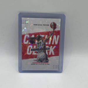 May include: A Caitlin Clark basketball trading card in a protective case. The card features a photo of Clark in action, with the words "Record Breaker" and "Indiana Fever" prominently displayed. The background is red and white.