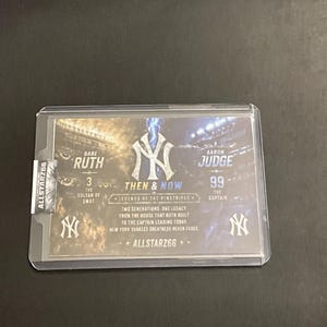 May include: A baseball trading card featuring Babe Ruth and Aaron Judge. The card is encased in a protective sleeve and includes the text "Then & Now" and "Legends of the Pinstripes". The card also displays the New York Yankees logo.
