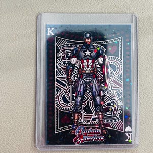 May include: A collectible playing card featuring Captain America in a blue, red, and white suit with a shield. The card has a holographic finish, the letter K in the corners, and the text Captain America at the bottom, all encased in a clear sleeve.