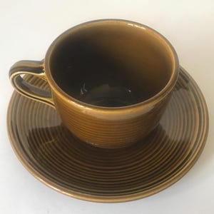 May include: A brown ceramic teacup and saucer set. The cup has a handle and a ridged texture, as does the matching saucer. The set is a warm, earthy brown color, suitable for serving tea or coffee.