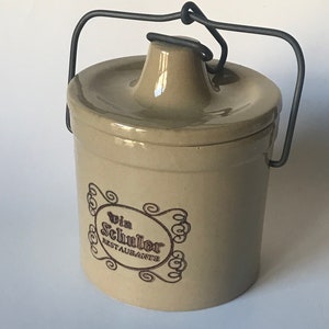 May include: Tan ceramic crock with a lid and a wire handle. The crock features a decorative oval logo with the text "Win Schuler Restaurants" in brown. The lid has a small spout. A vintage kitchenware item.