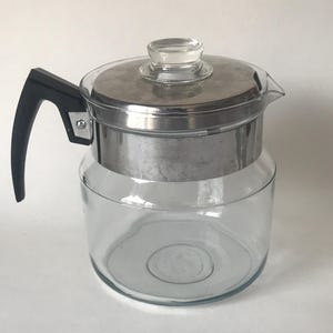 May include: A clear glass coffee pot with a silver lid and a black handle. The pot has a round body and a spout on the side.