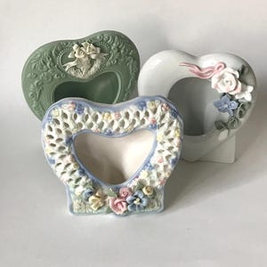 May include: Three heart-shaped ceramic picture frames. One is sage green with a floral design and a bow. Another is white with pink and blue flowers. The third is white with a blue and yellow floral design.