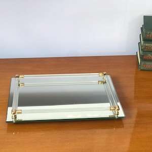 May include: A rectangular mirrored tray featuring a clear acrylic handle frame with polished gold-tone metal accents at each corner. The tray has a reflective glass base and a structured design with four vertical supports connecting the handles.