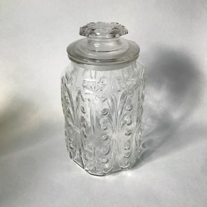 May include: A clear glass jar with a decorative lid. The jar has a textured, embossed pattern on the sides. The lid is round with a floral design on top. The jar is empty and sits on a white surface.