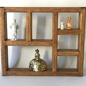May include: A wooden display shelf with five compartments, holding small figurines. The figurines include a white statue, a brass figure of a woman in a dress, and two other figurines. The shelf is made of light brown wood.
