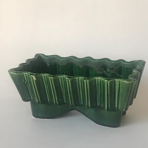 May include: A green ceramic planter with a wavy, scalloped rim and a unique, asymmetrical shape. The planter has a textured, ribbed design and stands on three legs.