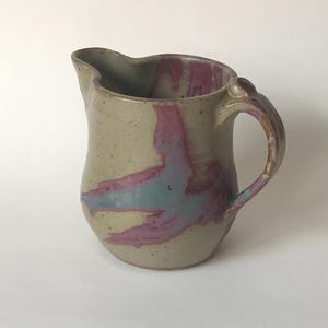 May include: A ceramic pitcher with a light brown glaze and pink and blue abstract designs. The pitcher has a rounded body and a handle.