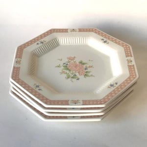 May include: A stack of four octagonal plates. Each plate has a white center with a floral design of pink roses and green leaves. The rim is bordered with a pink and white geometric pattern. The plates are stacked on top of each other.