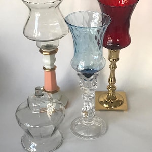 May include: A collection of decorative glass candle holders in various shapes and colors. Includes a clear glass jar with a lid, a clear glass candle holder with a pink and gold base, a blue textured glass candle holder, and a red glass candle holder.