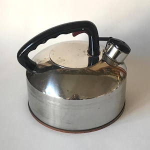 May include: A stainless steel stovetop kettle with a black handle and spout. The kettle has a round body and a black lid. The handle has a red button. The base of the kettle has a copper-colored ring.
