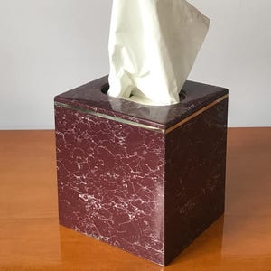 May include: A square tissue box with a burgundy marble pattern and a gold trim. White tissues are pulled from the top opening. The box is sitting on a wooden surface.