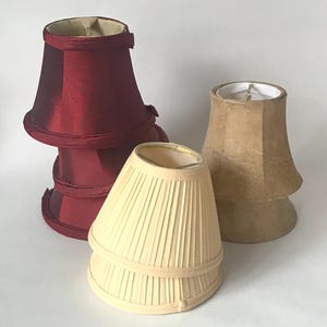 May include: Three lamp shades of varying styles and colors. One is a deep burgundy satin shade, another is a cream-colored pleated shade, and the third is a textured tan shade. Each shade features a flared base and a metal fitter ring inside.