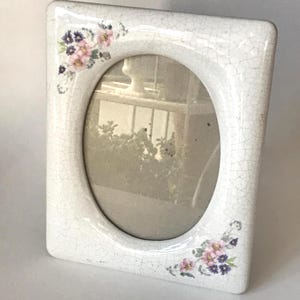 May include: White ceramic picture frame with an oval opening. The frame has a crackle finish and is decorated with clusters of pink and purple flowers in the corners. The frame is rectangular with rounded corners.