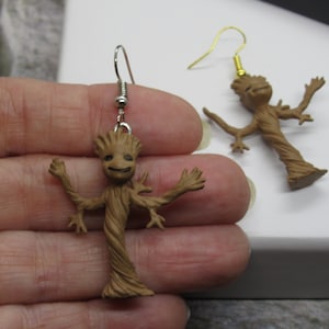 May include: A pair of earrings featuring a brown, tree-like character with outstretched arms. The character is reminiscent of the fictional character Groot from the Guardians of the Galaxy franchise.