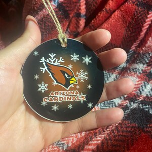 May include: A round, black and red Christmas ornament with a white snowflake design. The ornament features the Arizona Cardinals logo with the team name "Arizona Cardinals" printed below the logo.