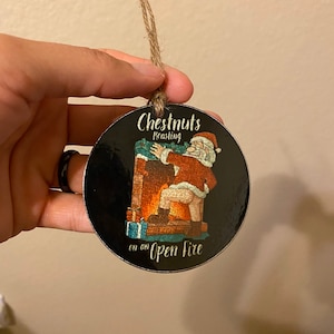 May include: A round, black Christmas ornament with a cartoon image of Santa Claus roasting chestnuts on an open fire. The ornament has the text "Chestnuts Roasting on an Open Fire" printed on it.