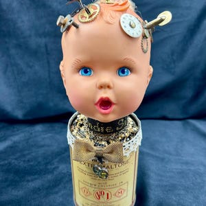 Peut inclure: Une tête de poupée vintage aux yeux bleus et la bouche ouverte est fixée à une boîte en métal avec un nœud doré et une étiquette qui indique "Extra Maltose" avec le texte "With Sodium Chloride 2% D No 1 M Mead Johnson & Co."