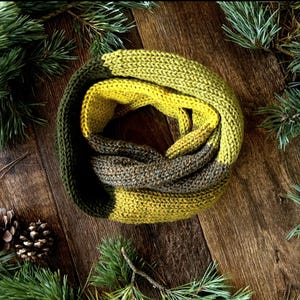 May include: A circular knit infinity scarf featuring a colour-blocked design with segments of dark forest green, bright chartreuse yellow, and a heathered grey-brown yarn, showcasing a consistent stockinette stitch pattern throughout the garment.