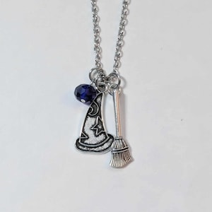 May include: A silver-toned necklace featuring a witch-themed charm set. The charms include a witch hat with a crescent moon and star design, a small broom, and a dark blue bead. The necklace hangs against a white background.