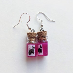 May include: A pair of novelty earrings featuring small glass bottles with cork stoppers. The bottles are filled with a vibrant pink liquid and have a small label with a black llama illustration. The earrings have silver-colored fish hook ear wires.