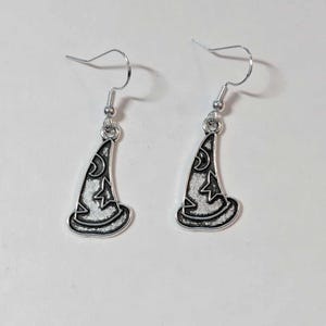 May include: A pair of silver-toned earrings featuring a witch hat design. The hats are outlined in black and filled with a white star and crescent moon pattern. The earrings hang from silver-colored ear wires.