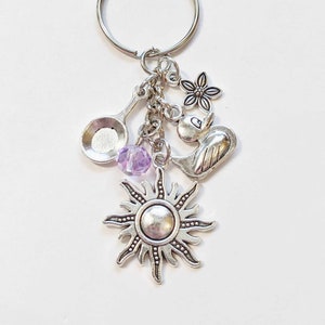 May include: Silver charm keychain with a sun, a bird, a flower, a spoon, and a purple bead.