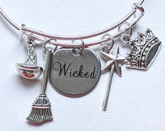 May include: Silver-toned bangle bracelet with multiple charms. Charms include a witch hat, broom, a round charm with the word "Wicked", a star wand, and a crown. The bracelet is on a white surface.