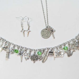 May include: A silver charm bracelet with green beads and charms featuring a pirate ship, a skull and crossbones flag, a fairy, a clock tower, a feather, a treasure chest, a crocodile, and a bow. The bracelet comes with a matching silver necklace with a charm that reads "All it takes is faith, trust & a little dust" and a pair of silver star-shaped earrings.