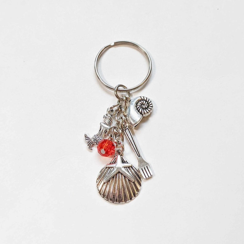 May include: Silver-coloured keyring with multiple charms, including a seashell, starfish, mermaid, and a red bead. The keyring is attached to a silver ring. The charms have a nautical theme.