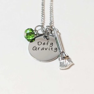 May include: Silver necklace with a round charm that says "Defy Gravity" and a small green crystal charm. The necklace also has a silver charm that looks like a broom.
