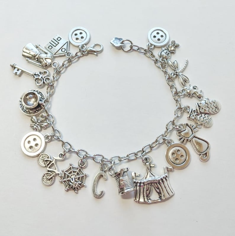 May include: A silver charm bracelet with various charms, including a house, a key, a teacup, a button, a spider web, a letter C, a spool of thread, a tent, a cat, a dragonfly, and a mitten.