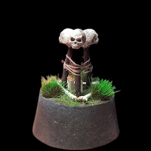 Op de afbeelding: Een handgemaakte miniatuur Halloween-sculptuur. Het heeft een donkergrijze basis met groen gras, een centrale structuur van houten palen en een schedel bovenop. De palen zijn met touw gebonden.