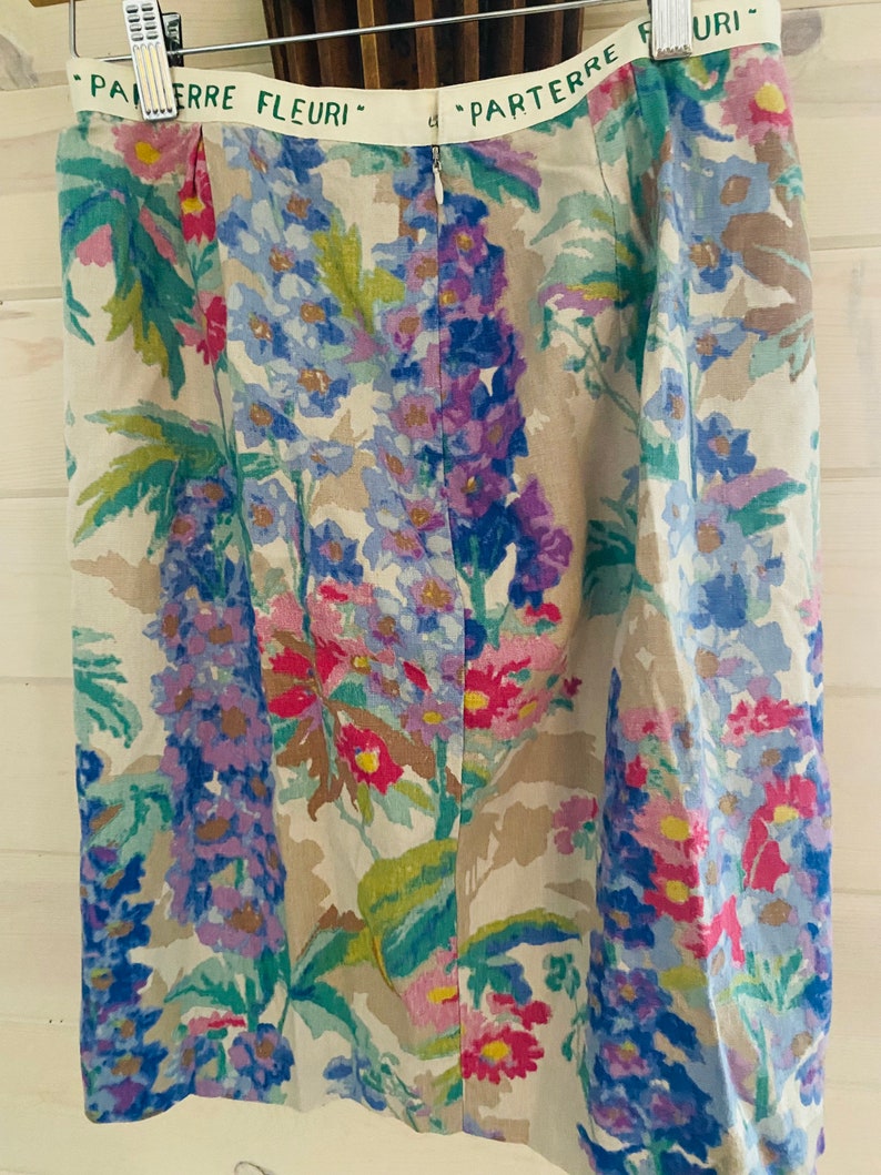 May include: A beige skirt with a floral print featuring pink, blue, and green flowers. The skirt has a zipper closure and a waistband with the text "Parterre Fleuri" in green.