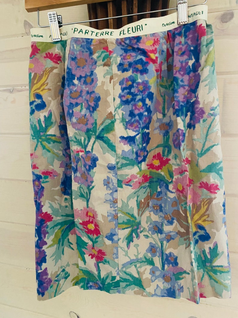 May include: A beige skirt with a floral print featuring blue, purple, pink, and green flowers. The skirt has a waistband with the text "PARTERRE FLEURI" in green.