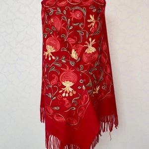 May include: A red shawl with embroidered floral designs in shades of gold, brown, and red. The shawl has fringe along the bottom edge.